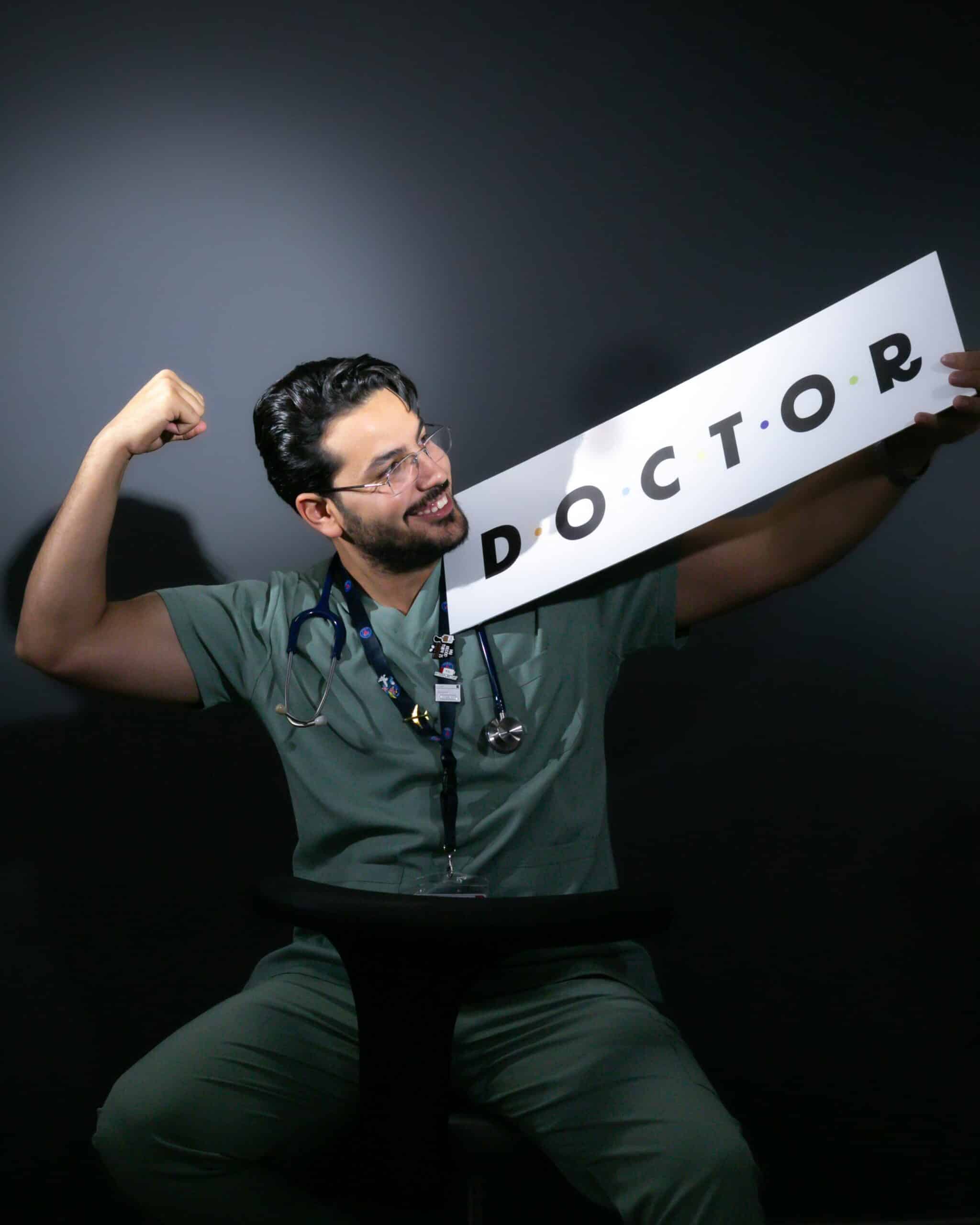 A doctor in scrubs dons a cheerful smile, holding a 'DOCTOR' sign, celebrating his profession.
