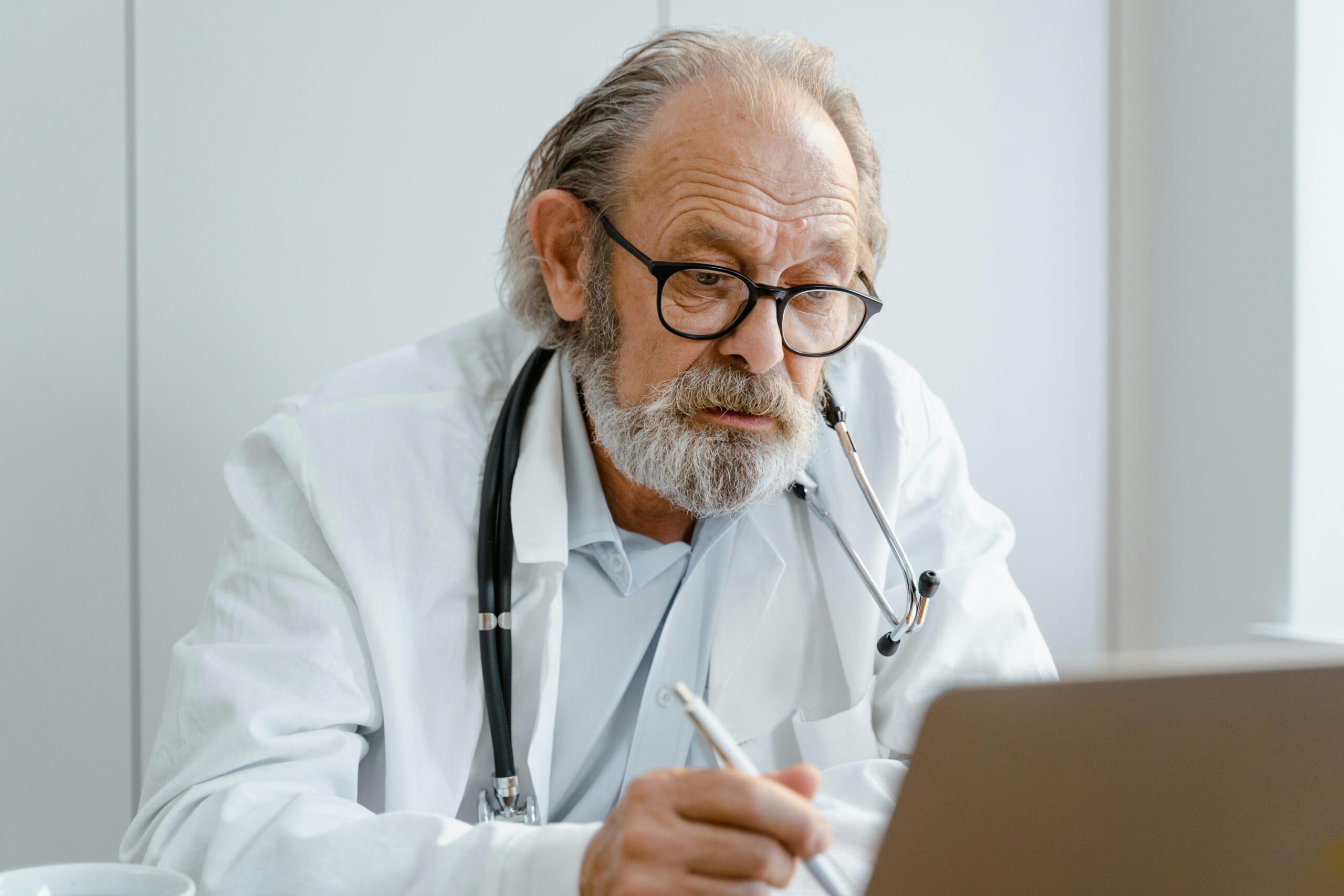 Elderly doctor with glasses and stethoscope working on a laptop in a medical office.
